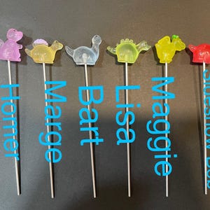 May include: Six colorful dinosaur-shaped cake toppers on metal skewers. The toppers are pink, yellow, blue, green, yellow, and red. The names Homer, Marge, Bart, Lisa, Maggie, and Sideshow Bob are written below each topper.