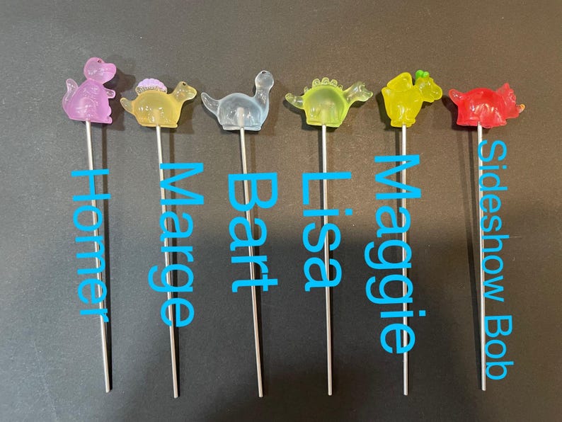 May include: Six colorful dinosaur-shaped cake toppers on metal skewers. The toppers are pink, yellow, blue, green, yellow, and red. The names Homer, Marge, Bart, Lisa, Maggie, and Sideshow Bob are written below each topper.