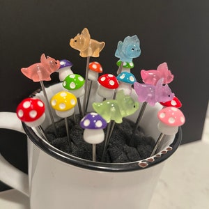 May include: A set of colorful mushroom and dinosaur shaped food picks in a white mug. The picks are made of glass and have a silver metal stem.