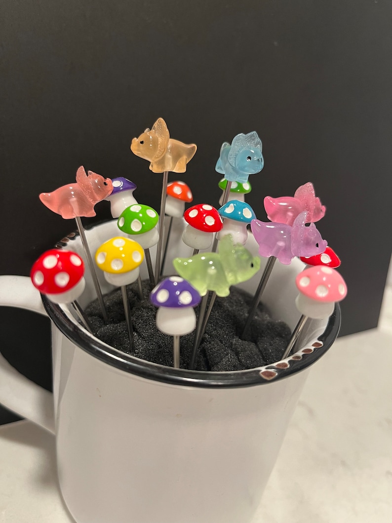 May include: A set of colorful mushroom and dinosaur shaped food picks in a white mug. The picks are made of glass and have a silver metal stem.