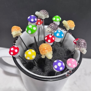 May include: A white ceramic mug with a black rim filled with colorful mushroom-shaped picks with tiny turtle figurines on top. The picks are arranged in a variety of colors, including red, green, yellow, purple, and pink.