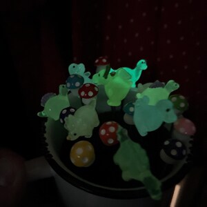 May include: A collection of glow-in-the-dark dinosaur and mushroom figurines in a white mug. The figurines are green, blue, and red.