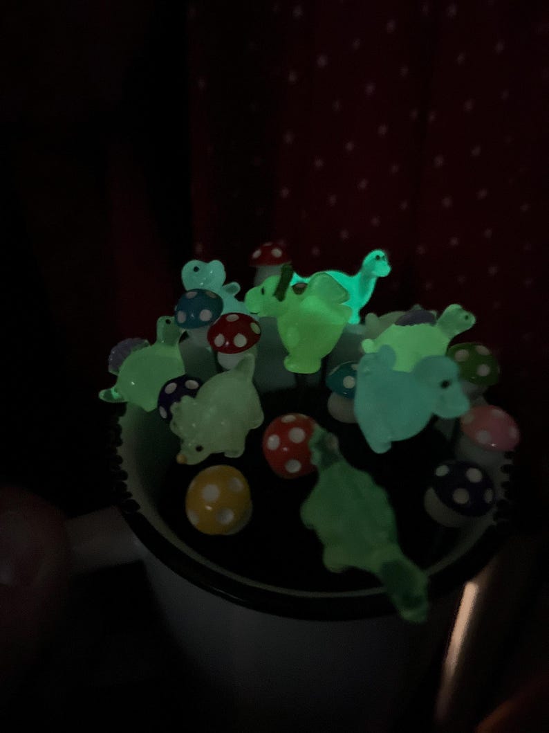 May include: A collection of glow-in-the-dark dinosaur and mushroom figurines in a white mug. The figurines are green, blue, and red.