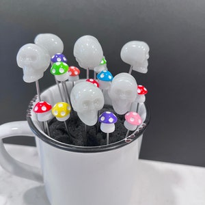 May include: A white ceramic mug filled with white skull-shaped cocktail picks topped with colorful mushroom-shaped toppers.