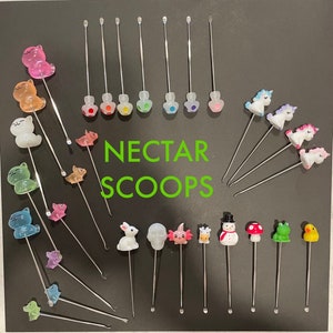 May include: A collection of nectar scoops with various colorful animal and character designs. The scoops have stainless steel stems and are topped with clear, decorative figures, including bears, unicorns, and a snowman. The text "NECTAR SCOOPS" is in green.