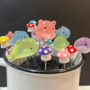 May include: A set of colorful glass animal and mushroom food picks. The picks feature a variety of designs, including dolphins, bears, and mushrooms with white polka dots. The picks are arranged in a white mug with a black rim.