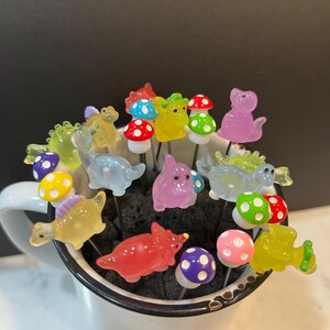 May include: A collection of colorful glass dinosaur figurines and mushroom-shaped toppers, arranged in a white mug. The figurines are translucent and feature various colors, including green, yellow, pink, blue, and red. The mushroom toppers have white tops with colorful polka dots.