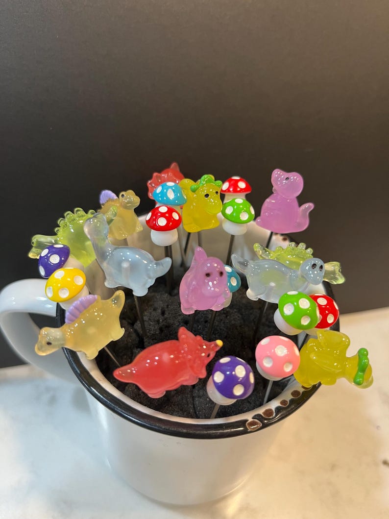 May include: A collection of colorful glass dinosaur figurines and mushroom-shaped toppers, arranged in a white mug. The figurines are translucent and feature various colors, including green, yellow, pink, blue, and red. The mushroom toppers have white tops with colorful polka dots.