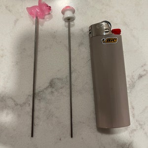 May include: Two metal skewers with pink and white ceramic toppers shaped like a pig and a mushroom. A gray lighter with the word "BIC" in yellow letters is also in the image.