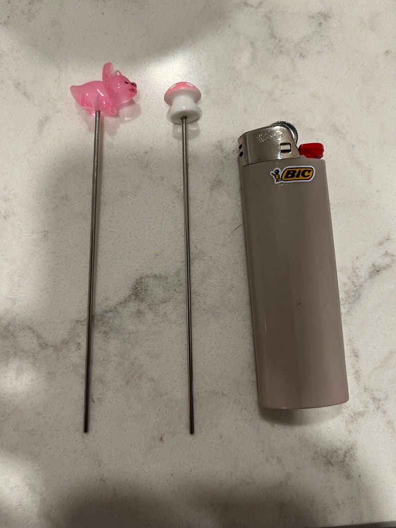 May include: Two metal skewers with pink and white ceramic toppers shaped like a pig and a mushroom. A gray lighter with the word "BIC" in yellow letters is also in the image.