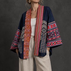 Peut inclure: Une veste bleu foncé ouverte sur le devant avec des manches larges et un motif patchwork. Le vêtement présente des panneaux de broderies géométriques complexes rouges, orange et blanches, ainsi que des sections de tissu à carreaux bleu et marron.