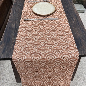 May include: A rectangular table runner featuring a repeating rust-coloured and cream fan-shaped wave pattern. The fabric is laid flat across a dark wooden table, with a single round ceramic plate and a pair of metal chopsticks placed on top.