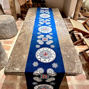 Puede incluir: Un camino de mesa azul largo y rectangular con un patrón repetitivo de diseños geométricos blancos y rojos. El camino de mesa está colocado sobre una mesa de piedra, agregando un toque decorativo. El diseño incluye motivos circulares y cuadrados.