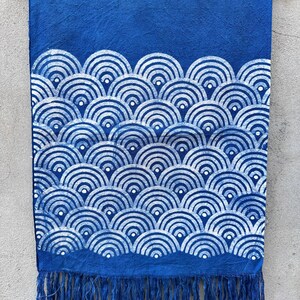 K&ouml;nnte beinhalten: Blauer Stoff mit wei&szlig;em, wellenartigem Muster und Fransen. Der Stoff h&auml;ngt an einem Holzb&uuml;gel. Das Design zeigt sich wiederholende Halbkreise, die einen strukturierten, dekorativen Effekt erzeugen. Der untere Rand hat Fransen.