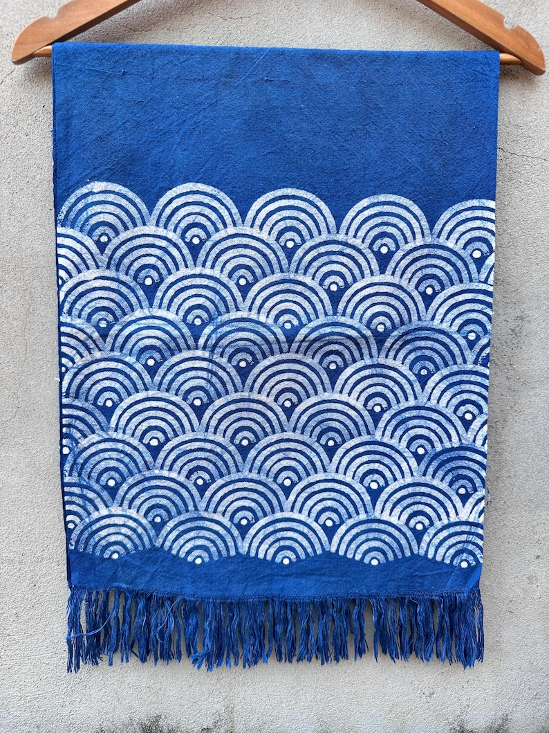 K&ouml;nnte beinhalten: Blauer Stoff mit wei&szlig;em, wellenartigem Muster und Fransen. Der Stoff h&auml;ngt an einem Holzb&uuml;gel. Das Design zeigt sich wiederholende Halbkreise, die einen strukturierten, dekorativen Effekt erzeugen. Der untere Rand hat Fransen.