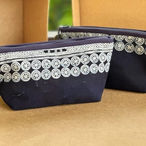 Könnte beinhalten: Zwei marineblaue Kosmetiktaschen mit weißen, gestickten Radmustern. Die Taschen haben einen Reißverschluss und eine Quaste mit einem goldenen Anhänger. Die Taschen befinden sich in einer braunen Box.