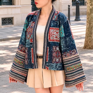 May include: A long-sleeved open-front jacket featuring a patchwork design with deep blue, red, and teal floral and geometric patterns. The garment includes wide sleeves and a dark trim with a repeating white cross motif along the front edges.