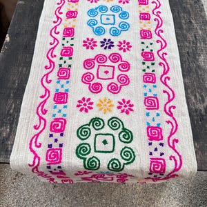 以下が含まれることがあります： 鮮やかな多色の刺繍パターンが施された、オフホワイトの長方形のテーブルランナー。ピンク、青、緑、黄色、紫の幾何学模様、渦巻き、花柄が垂直の列に並び、繰り返し配置されているデザインが特徴です。
