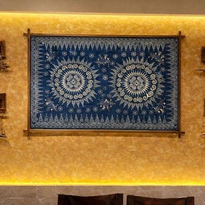 Könnte beinhalten: Eine Wanddekoration mit einem großen, gerahmten blauen Textil mit weißen kreisförmigen Mustern und einem geometrischen Rand. Kleinere, holzgerahmte Webstücke sind an der strukturierten, goldfarbenen Wand angebracht.