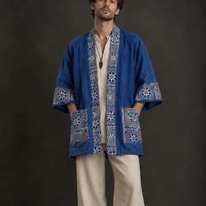 May include: A blue open-front jacket with wide sleeves and two front pockets. The garment features a patterned trim along the collar and sleeve cuffs, displaying white star-like motifs on a blue background. It is worn over a beige shirt and trousers.