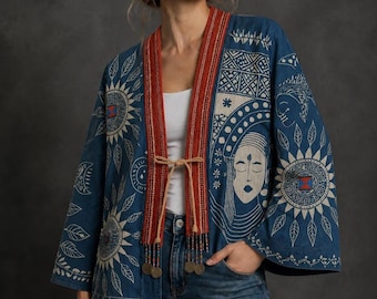 May include: A blue open-front jacket featuring white line art of suns, leaves, and a stylized face. The garment has a wide, textured rust-colored trim along the front opening, secured with thin tan ties that feature small dangling metal coin charms.