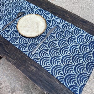 以下が含まれることがあります： 濃い木製のテーブルの上に、青と白の模様のテーブルランナー、お皿、フォーク、ナイフが置かれています。 ランナーは繰り返しの波のデザインが特徴です。 お皿はベージュで茶色の縁があり、カトラリーは銀色です。