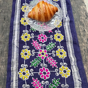 May include: A golden-brown croissant sits on a clear, textured glass plate placed atop a dark blue table runner. The runner features a repeating pattern of embroidered yellow flowers, pink blossoms, and green leaves with white striped borders.