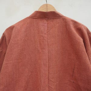 Puede incluir: Chaqueta de manga larga color &oacute;xido con cuello mandar&iacute;n, colgada de una percha de madera. La parte trasera de la chaqueta es visible, mostrando la textura de la tela y las costuras a medida. La prenda parece estar hecha de un material ligero.