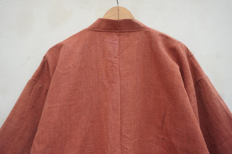 Puede incluir: Chaqueta de manga larga color &oacute;xido con cuello mandar&iacute;n, colgada de una percha de madera. La parte trasera de la chaqueta es visible, mostrando la textura de la tela y las costuras a medida. La prenda parece estar hecha de un material ligero.