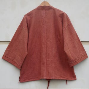 Puede incluir: Chaqueta de color &oacute;xido, de manga corta, con cuello mandar&iacute;n, colgada de una percha de madera. La chaqueta tiene un corte holgado y un cord&oacute;n en la parte inferior. La tela parece ser una mezcla de algod&oacute;n o lino texturizado.