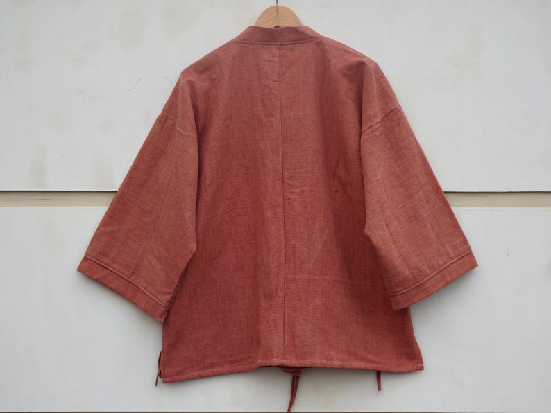 Puede incluir: Chaqueta de color &oacute;xido, de manga corta, con cuello mandar&iacute;n, colgada de una percha de madera. La chaqueta tiene un corte holgado y un cord&oacute;n en la parte inferior. La tela parece ser una mezcla de algod&oacute;n o lino texturizado.