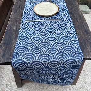 May include: A blue and white patterned table runner with a repeating wave design. A beige plate and cutlery are placed on top of the runner, which is laid out on a dark wooden table. The runner adds a decorative touch to the table setting.