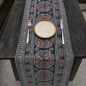May include: A navy blue table runner with a repeating red and beige floral pattern. A beige plate, fork, and knife are set on the runner. The runner is on a dark wooden table.