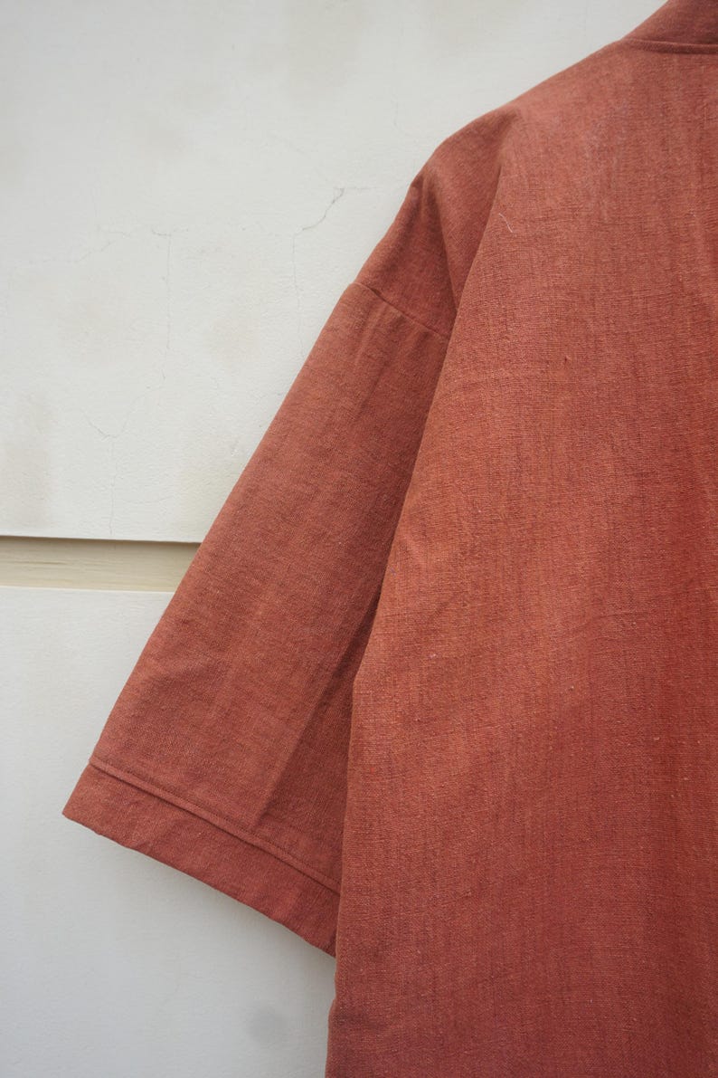Puede incluir: Primer plano de una camisa de manga corta de color &oacute;xido. La tela tiene un aspecto texturizado y la manga est&aacute; doblada en el pu&ntilde;o. La camisa est&aacute; hecha de un material suave y tiene un ajuste relajado. El fondo es una pared lisa de color blanquecino.