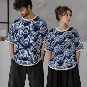 Puede incluir: Dos camisetas de manga corta y cuello redondo con un patrón repetitivo de semicírculos concéntricos azules y blancos con pequeños puntos rojos. Las camisetas tienen un borde de color beige claro en el cuello y el dobladillo, con pantalones negros.
