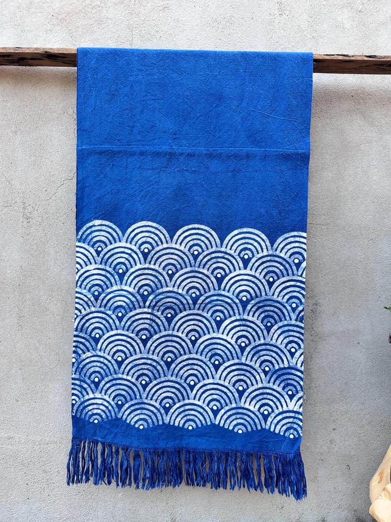 K&ouml;nnte beinhalten: Ein leuchtend blaues Textil mit wei&szlig;em Wellenmuster und Fransen. Der Stoff ist ein tiefes Blau mit einem sich wiederholenden Design aus wei&szlig;en, halbkreisf&ouml;rmigen Wellen. Der untere Rand hat Fransen.
