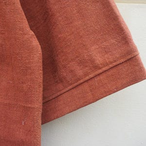 Puede incluir: Primer plano de una secci&oacute;n de tela de color &oacute;xido, posiblemente lino o un material tejido similar. La tela tiene un aspecto texturizado con hilos visibles y un brillo sutil. El borde de la tela est&aacute; cuidadosamente doblado y cosido.