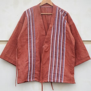 Puede incluir: Chaqueta estilo kimono de color &oacute;xido con detalles de rayas verticales en blanco, morado y marr&oacute;n. La chaqueta tiene cuello en V y mangas cortas, con lazos en la cintura. Cuelga de una percha de madera.