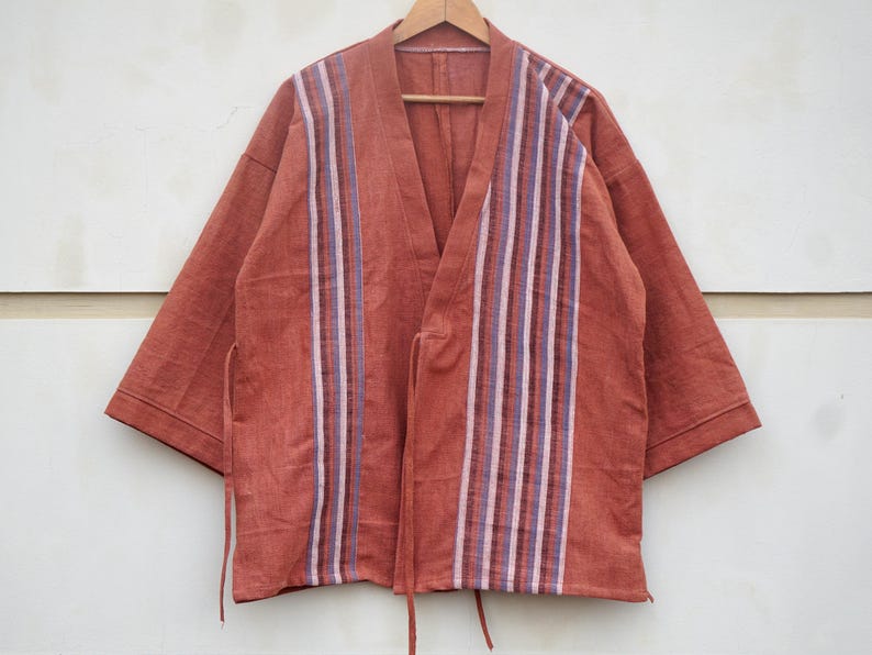Puede incluir: Chaqueta estilo kimono de color &oacute;xido con detalles de rayas verticales en blanco, morado y marr&oacute;n. La chaqueta tiene cuello en V y mangas cortas, con lazos en la cintura. Cuelga de una percha de madera.