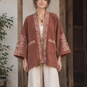 Puede incluir: Una chaqueta de color óxido con frente abierto, mangas anchas y un borde decorativo con patrones geométricos en tonos rosa, morado y dorado. La prenda incluye dos bolsillos delanteros y se lleva sobre una blusa blanca lisa y pantalones claros.