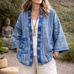 May include: A blue and white open-front jacket featuring a repeating geometric and floral pattern, wide three-quarter length sleeves, and a single patch pocket on the front. The garment is worn over a plain white top and includes a long beaded necklace.