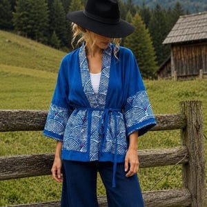 Könnte beinhalten: Eine blaue Jacke im Kimono-Stil mit einem weißen fächerförmigen Muster und kleinen violetten Akzenten. Das Kleidungsstück hat weite Ärmel, ein Wickeldesign mit einem passenden Bindegürtel aus Stoff und ein weißes Unterhemd unter der Vorderseite.