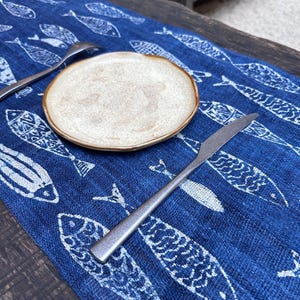 Peut inclure: Une petite assiette ronde beige avec un bord marron est posée sur une nappe bleue à motifs de poissons blancs. Un couteau et une cuillère argentés sont également sur la table. La table est sur une surface en bois.