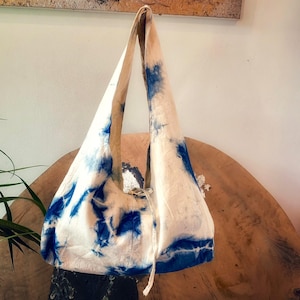 May include: A white tote bag with a blue tie-dye pattern. The bag has a triangular shape with long straps and a drawstring closure. The bag is hanging on a wooden surface.