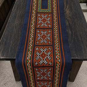 May include: A long, rectangular dark blue table runner featuring intricate cross-stitch embroidery. The design includes repeating geometric square patterns in orange, white, and green, framed by vertical stripes of the same vibrant colors.