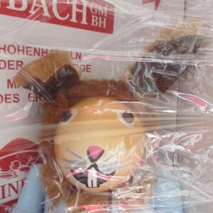 May include: A wooden toy in the shape of a rabbit, with a light brown head, long ears, and a pink nose. The rabbit is wearing a light blue outfit with orange and yellow accents. The toy is wrapped in clear plastic.