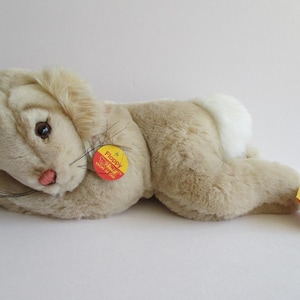 May include: A beige floppy rabbit plush toy laying on its side with its head resting on its front paws. The toy has a white patch on its back and a yellow tag that says "Floppy" on it.