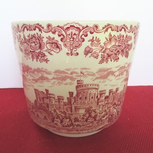 May include: A cream-colored ceramic planter featuring a detailed red transferware pattern. The design displays a large castle with turrets and a flag, surrounded by trees and landscape, topped with a decorative floral border around the rim.