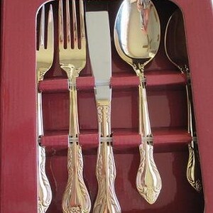 May include: A set of gold-colored silverware, including forks, knives, and spoons, arranged in a red box. The silverware has ornate handles and a shiny finish, suggesting a formal dining set.