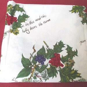 May include: White tablecloth with a Christmas holly and berry design. The design includes green holly leaves, red berries, and purple grapes. The text "tree in the wood the holly bears the corona" is written in cursive.